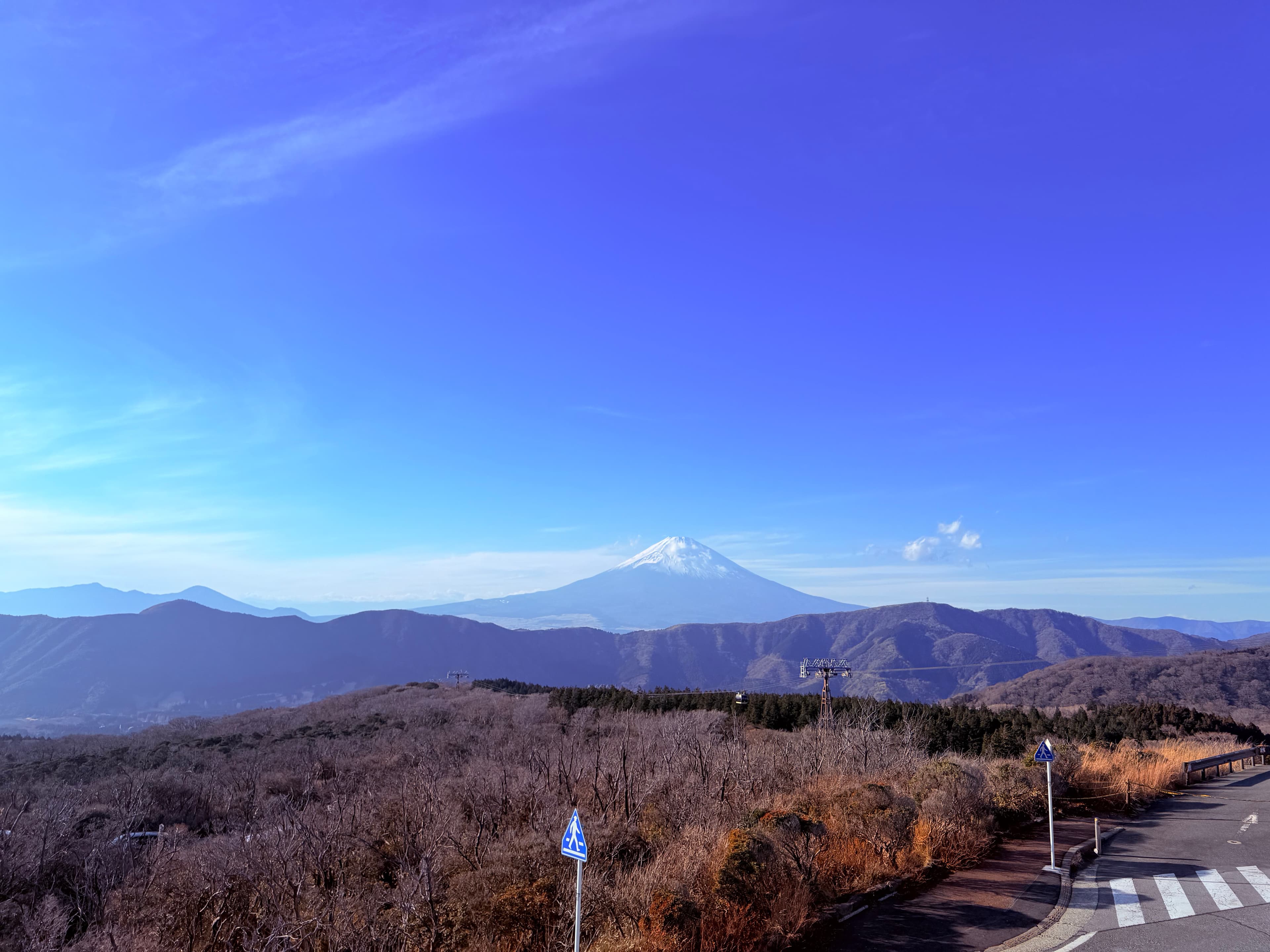 Snow-capped Mount Fuji rises above a mountain range and brown hills under clear blue skies.