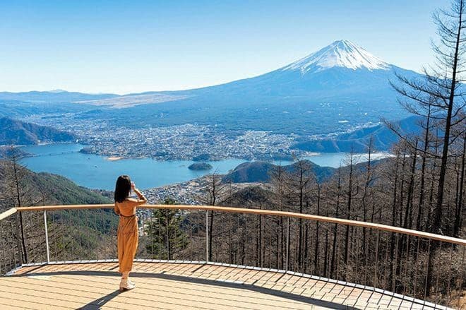 Woman on a wooden deck overlooking snow-capped Mount Fuji and a blue lake below.