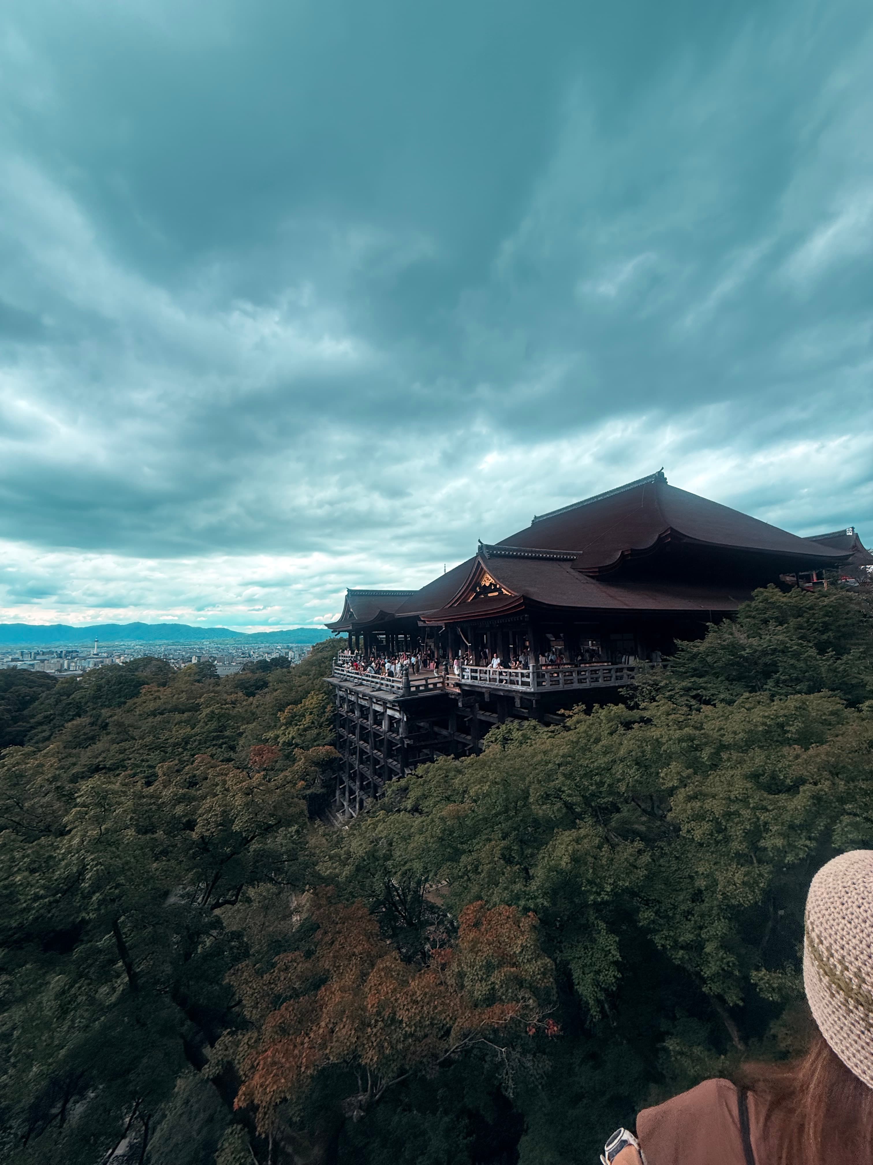 Kiyomizu-dera temple's wooden stage overlooks a lush forest and city under dramatic cloudy skies.
