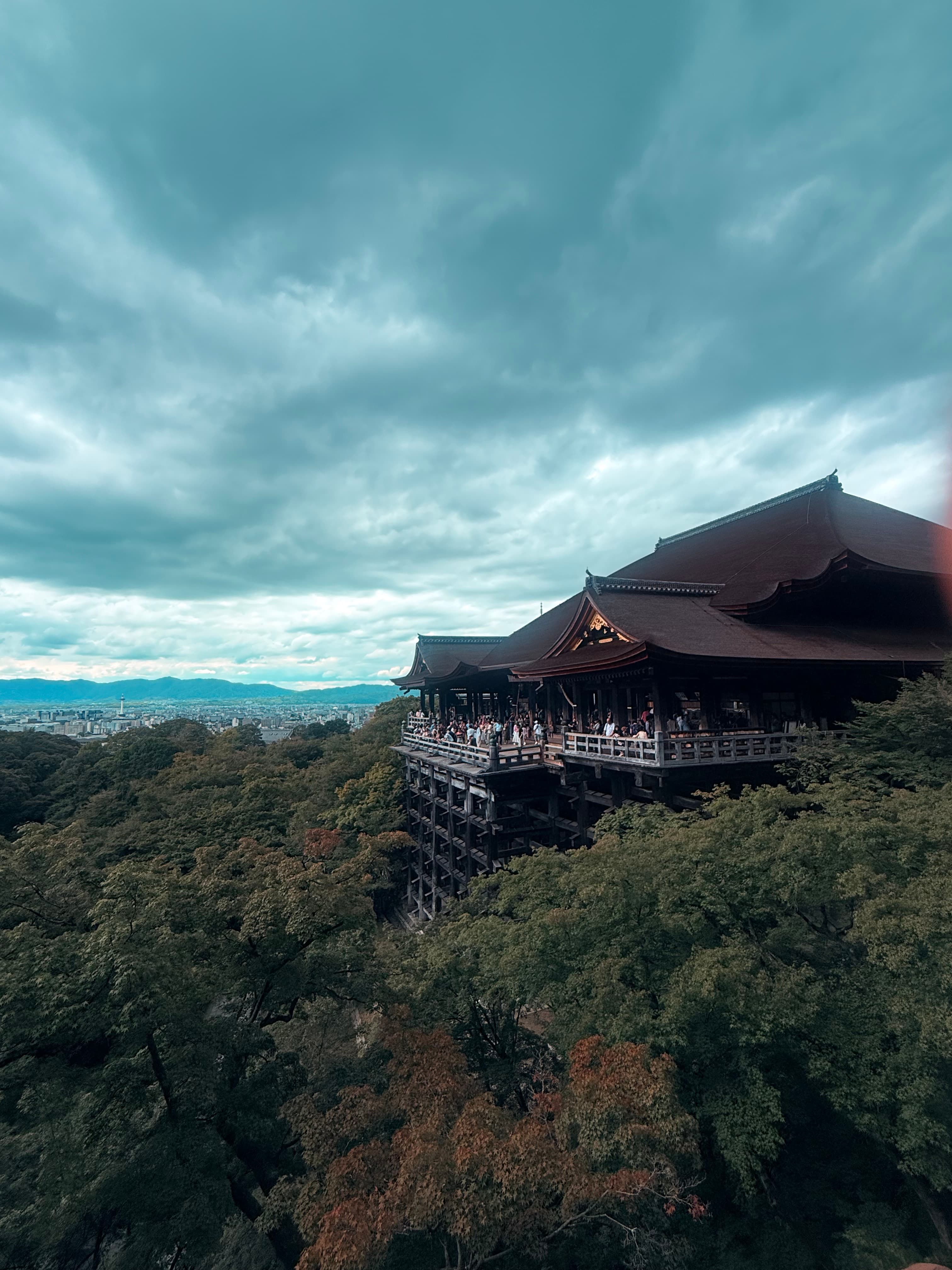 Kiyomizu-dera temple's wooden stage overlooks a lush forest and city under dramatic, overcast skies.