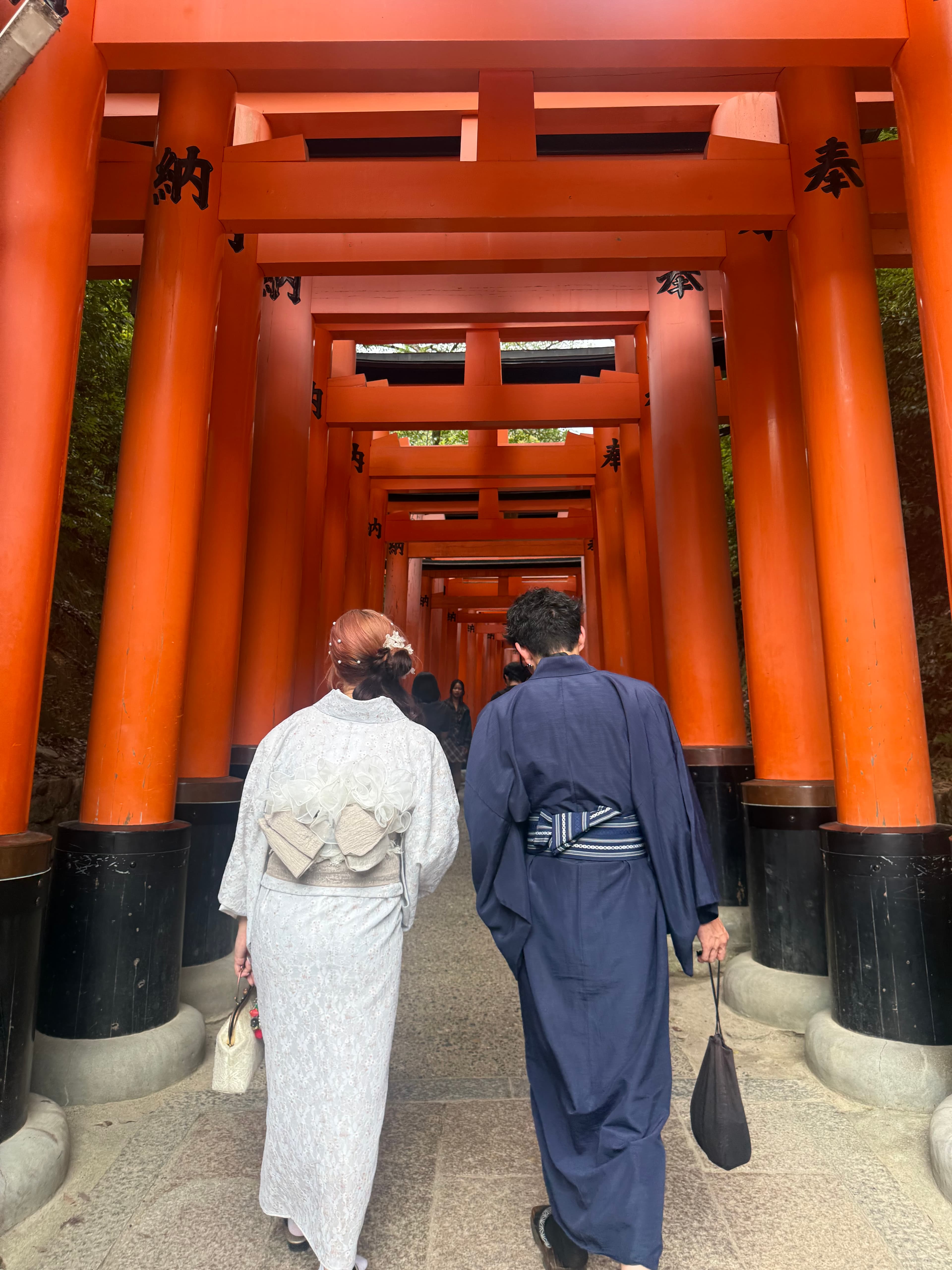 A couple wearing traditional kimonos walks through a path of vibrant orange torii gates.